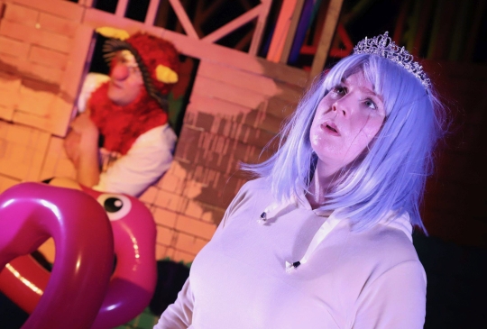 Lisa-Marie Gerl, eine weiblich gelesene weiße Schauspielerin, steht im rechten vorderen Bereich des Bildes. Sie trägt eine blaue Perücke, eine Krone und einen weißen Pullover. Im Hintergrund sieht man die Bühne mit pinken Schwimmeifen in Form von Flamingos. Aus einem Fenster in der Bühnenwand im Hintergrund guckt ein männlich gelesener, weißer Schauspieler mit roter Clownsnase und einer Mütze mit Hörnern.