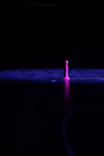 Auf einem schwarzen Bühnenboden ist ein pinker Dildo in einer Nahaufnahme zu sehen, der von rechts leicht beleuchtet wird. Der Rest des Bühnenraums ist dunkel.